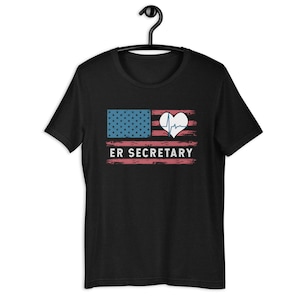 May include: Black t-shirt featuring a distressed American flag design with a heart-shaped EKG and the text "ER SECRETARY" in white. The flag has blue stars and red stripes. The shirt is hanging on a black hanger.