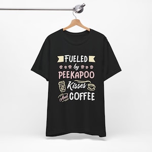Può includere: T-shirt nero con la scritta "Fueled by Peekapoo Kisses and Coffee" in bianco, rosa e giallo. Il design include impronte di zampe, una tazza di caffè e un'icona di tazza di caffè.