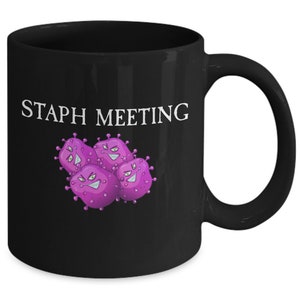 May include: Black coffee mug with the text "STAPH MEETING" in white lettering and a cartoon image of purple bacteria with angry faces.