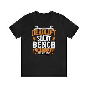 Może przedstawiać: Czarny T-shirt z białym tekstem, który brzmi "Deadlift Squat Bench 1000 LB Club Est. Date Here" w stylu vintage. Tekst jest otoczony pomarańczowymi symbolami podnoszenia ciężarów.