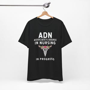 May include: Black t-shirt with the text "ADN Associate's Degree in Nursing In Progress". The design includes a floral caduceus symbol in pink, white, and green.