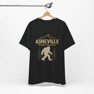 May include: Black t-shirt with a graphic featuring the text "Asheville North Carolina" above a Bigfoot silhouette and pine trees. A mountain range with a cyclist is above the text.