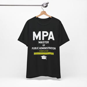 Puede incluir: Camiseta negra con el texto blanco "MPA MASTER OF PUBLIC ADMINISTRATION COMPLETED" y un gráfico de birrete. La camiseta está colgada en una percha de madera.