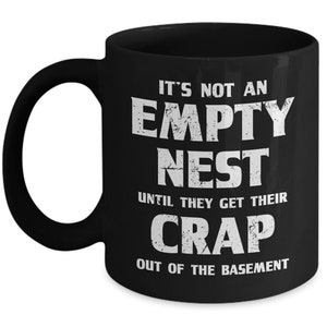Pode incluir: Caneca de caf&eacute; de cer&acirc;mica preta com texto branco. O texto na caneca diz: "IT'S NOT AN EMPTY NEST UNTIL THEY GET THEIR CRAP OUT OF THE BASEMENT". A caneca tem uma al&ccedil;a grande e um acabamento brilhante.