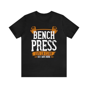 Peut inclure: T-shirt noir avec un motif blanc et orange. Le motif affiche "BENCH PRESS 225 LB CLUB" avec une illustration d'haltère et "EST. DATE HERE". Un t-shirt décontracté pour les amateurs d'haltérophilie.