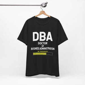 May include: Black t-shirt with the white text "DBA DOCTOR OF BUSINESS ADMINISTRATION IN PROGRESS" and a yellow progress bar graphic. The shirt is hanging on a wooden hanger.