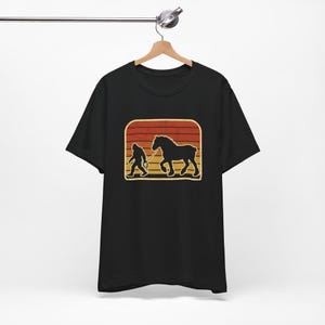 Könnte beinhalten: Schwarzes T-Shirt mit einem Retro-Grafikdesign. Das Design zeigt eine Silhouette einer Person, die ein Pferd führt, vor einem Hintergrund aus orangefarbenen und gelben horizontalen Streifen.