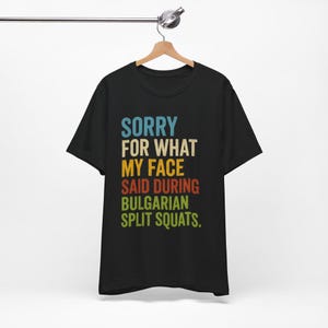 Pode incluir: Uma t-shirt preta de manga curta com o texto multicolorido SORRY FOR WHAT MY FACE SAID DURING BULGARIAN SPLIT SQUATS. As letras estão dispostas em linhas empilhadas com tons de azul, bege, amarelo, laranja e verde na parte frontal.