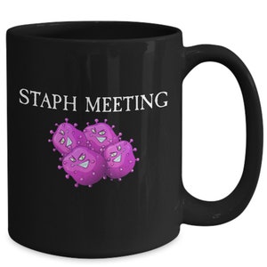 May include: Black coffee mug with the text "STAPH MEETING" in white lettering. The mug features a cartoon image of four purple bacteria with angry faces.