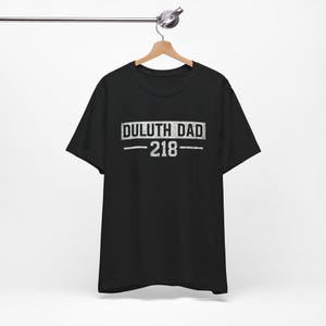 May include: A black short-sleeved t-shirt featuring white distressed-style text on the chest that reads DULUTH DAD with the number 218 centered below it between two horizontal lines. The garment is displayed on a wooden hanger.
