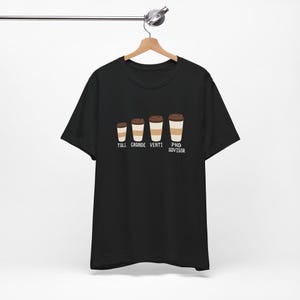 May include: A black short-sleeved t-shirt featuring a graphic of four coffee cups in increasing sizes. Below each cup is text reading TALL, GRANDE, VENTI, and PHD ADVISOR. The design is centred on the chest of the shirt, which hangs on a wooden hanger.