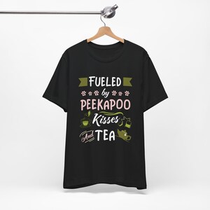 Può includere: T-shirt nera con la scritta "Fueled by Peekapoo Kisses and Tea" in bianco, rosa e verde. Il design include impronte di zampe, una tazza da tè e una teiera. La maglietta è appesa a una gruccia di legno.