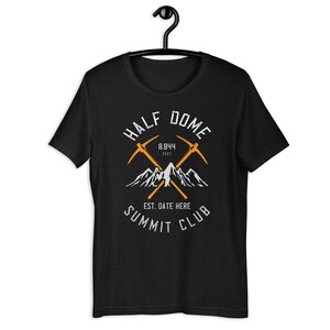 May include: Black t-shirt with a graphic featuring the words "HALF DOME" and "SUMMIT CLUB". The design includes crossed orange ice axes, mountain silhouettes, and the text "8,844 FEET". The shirt is hanging on a black hanger.