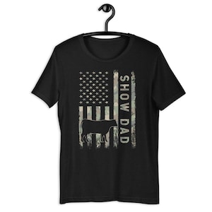 May include: Black t-shirt with a camouflage American flag design and the text "Show Dad" printed vertically on the right side of the flag. A silhouette of a cow is in front of the flag.