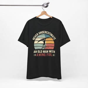 May include: Black t-shirt featuring a vintage-style graphic that reads "Never underestimate an old man with a wing foil". The graphic depicts a silhouette of a person holding a wing foil against a sunset backdrop.