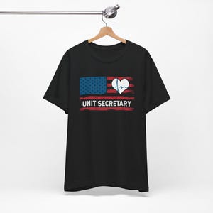 May include: Black t-shirt with a graphic featuring a stylized American flag design. The design includes a blue section with white stars, red stripes, a white heart with a heartbeat line, and the text "UNIT SECRETARY" in white.