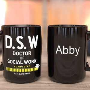 May include: Two black mugs with white text. One mug says "D.S.W. Doctor of Social Work Completed" with a yellow progress bar and the text "Est. Date Here". The other mug says "Abby".
