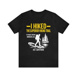 May include: Black t-shirt with a white and yellow graphic. The graphic reads "-I HIKED- THE SUPERIOR HIKING TRAIL 310 MILES / 500 KM MINNESOTA, USA" with a hiker silhouette and trees. The text "EST. DATE HERE" is also included.