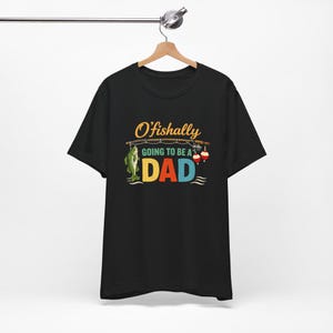 May include: Black t-shirt with the text "O'fishally Going to be a Dad" in orange, yellow, blue, and green. The design includes a cartoon fish and fishing bobbers. The shirt is hanging on a wooden hanger.