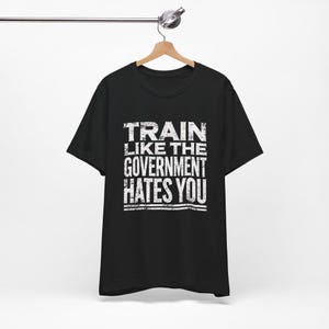 Peut inclure: Un t-shirt noir à manches courtes avec un texte blanc au style vieilli sur la poitrine qui indique : TRAIN LIKE THE GOVERNMENT HATES YOU. Le t-shirt est présenté sur un cintre en bois suspendu à une barre métallique sur fond blanc.