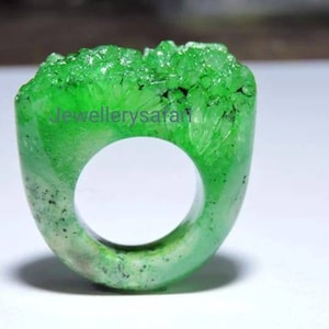 Könnte beinhalten: Ein grüner Ring aus durchsichtigem Material mit rauer, kristalliner Textur. Der Ring hat die Form eines abgerundeten Rechtecks mit einer großen Öffnung in der Mitte.