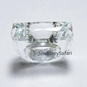Puede incluir: Un anillo de cristal transparente con una piedra de forma cuadrada. El anillo es de metal y tiene un acabado pulido.