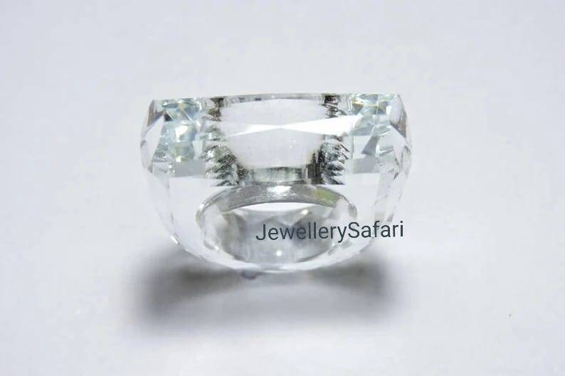 Puede incluir: Un anillo de cristal transparente con una piedra de forma cuadrada. El anillo es de metal y tiene un acabado pulido.