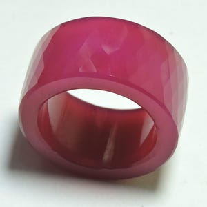 Könnte beinhalten: Ein breiter, facettierter Ring in leuchtendem Magenta. Der Ring hat ein dickes Band und eine geometrische, strukturierte Oberfläche. Die polierte Oberfläche reflektiert das Licht und erzeugt ein Schattenspiel. Dieser auffällige Ring ist ein auffälliges Schmuckstück.