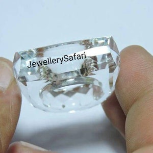 Puede incluir: Un anillo de cristal transparente con un dise&ntilde;o cuadrado facetado. El anillo est&aacute; sostenido por una mano y el texto "JewellerySafari" es visible en el anillo.