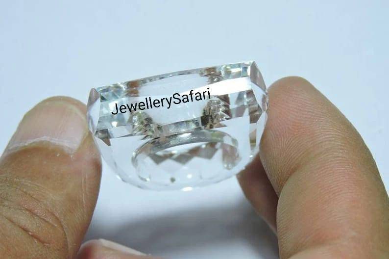 Puede incluir: Un anillo de cristal transparente con un dise&ntilde;o cuadrado facetado. El anillo est&aacute; sostenido por una mano y el texto "JewellerySafari" es visible en el anillo.