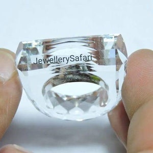 Puede incluir: Un anillo de cristal transparente con un dise&ntilde;o facetado. El anillo se sostiene en una mano y el texto "JewellerySafari" es visible en el anillo.