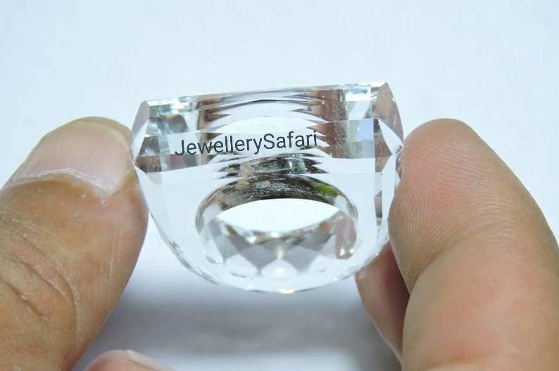 Puede incluir: Un anillo de cristal transparente con un dise&ntilde;o facetado. El anillo se sostiene en una mano y el texto "JewellerySafari" es visible en el anillo.