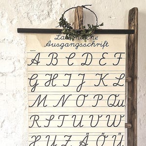 Può includere: Un grafico dell'alfabeto in stile vintage con il titolo tedesco "Lahnische Ausgangsschrift" in elegante corsivo. Il grafico presenta le lettere dalla A alla Ü e numeri romani. Il grafico è montato su una scala di legno.