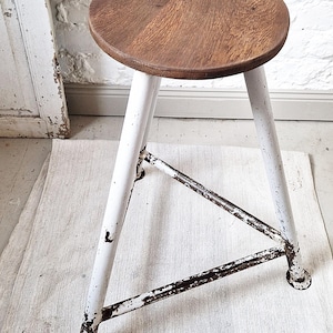 Könnte beinhalten: Vintage Holzstuhl mit runder, brauner Sitzfläche und weißen Metallbeinen. Die Beine sind durch Metallstreben verbunden und weisen Gebrauchsspuren auf. Der Hocker steht auf einem weißen Teppich vor einer weißen Backsteinmauer.