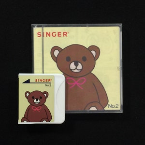 May include: A white plastic sewing machine embroidery card with a brown teddy bear wearing a pink bow. The card has the text "SINGER No. 2" printed on it. The card is next to a clear plastic case with a yellow background and a larger brown teddy bear wearing a pink bow. The case has the text "SINGER" printed on it. The case also has the text "No. 2" printed on it.