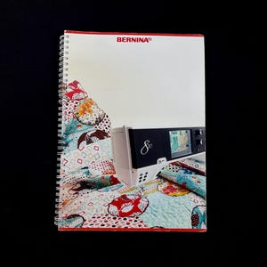 Könnte beinhalten: Ein spiralgebundenes Bernina-Nähmaschinenhandbuch mit weißem Einband und dem Markennamen in Rot. Das Handbuch ist auf einer Seite mit einem farbenfrohen Quiltmuster geöffnet. Eine weiß-schwarze Nähmaschine ist sichtbar.