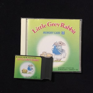 May include: A black memory card with the text "MEMORY CARD CHARACTER SERIES LITTLE GREY RABBIT" and a picture of a rabbit in a blue dress. The card is next to a CD case with the text "Little Grey Rabbit MEMORY CARD 7" and a picture of a rabbit in a blue dress.
