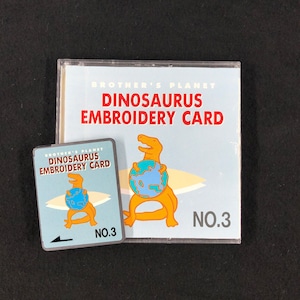 May include: Brother's Planet Dinosaur Embroidery Card No. 3. A blue and white card with a cartoon dinosaur holding a globe. The card is packaged in a clear plastic sleeve.