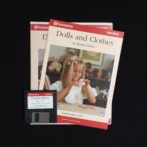 May include: A set of two embroidery disks for a sewing machine. The disks are labelled "Dolls and Clothes by Martha Pullen" and "Embroidery 115". The disks are packaged in a clear plastic sleeve with a white background. The image shows a young girl holding a doll and a teddy bear.