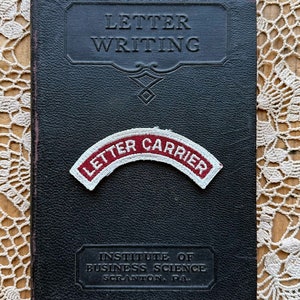 May include: A black leather-bound book titled "Letter Writing" with a red and white patch that reads "Letter Carrier" on the front cover. The book is from the Institute of Business Science in Scranton, PA.