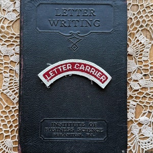 May include: A black leather-bound book titled "Letter Writing" with a red and white "Letter Carrier" patch on the front cover. The book is on a white crocheted doily. The book also has the text "Institute of Business Science Scranton, Pa."