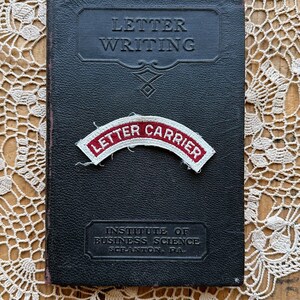 May include: A black leather-bound book titled "Letter Writing" with a red and white "Letter Carrier" patch on the front cover. The book is on a white crocheted doily. The book also says "Institute of Business Science Scranton, PA."
