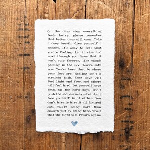 May include: A white paper with printed text and a small blue heart illustration. The text offers words of encouragement and support, set against a rustic wooden background. The paper has a deckled edge.