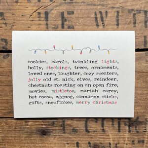 Op de afbeelding: Een witte kaart met een lijst van kersttradities geschreven in kleurrijke tekst. De tekst luidt: "koekjes, kerstliederen, twinkelende lichtjes, hulst, kousen, bomen, kerstversiering, dierbaren, gelach, gezellige truien, de oude kerstman, elven, rendieren, kastanien die aan het open vuur geroosterd worden, films, mistletoe, Mariah Carey, warme chocolademelk, advocaat, kaneelstokjes, cadeaus, sneeuwvlokken, vrolijk kerstfeest".