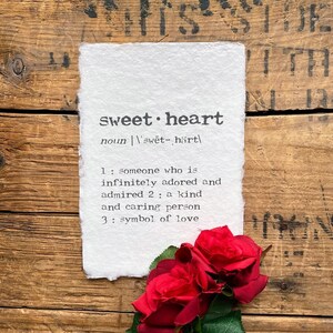 May include: A white paper with the word "sweetheart" written in black text, with a definition of the word below. The paper is sitting on a wooden surface with red roses in the bottom right corner.