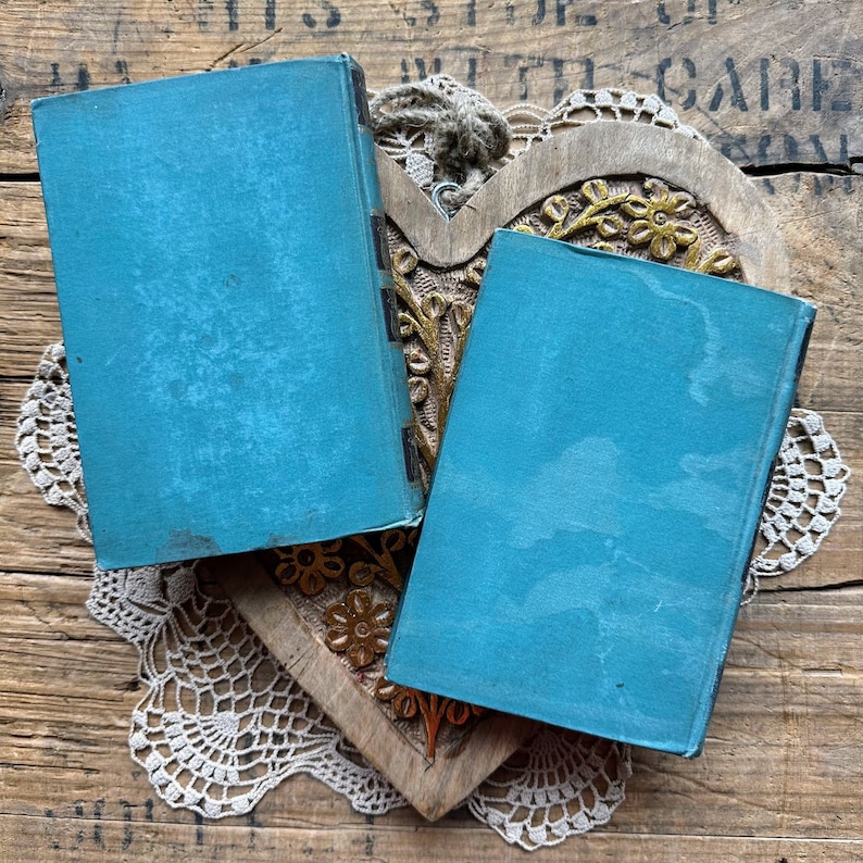 May include: Two vintage blue hardback books with gold accents on the spine, resting on a wooden heart-shaped object with intricate carvings.