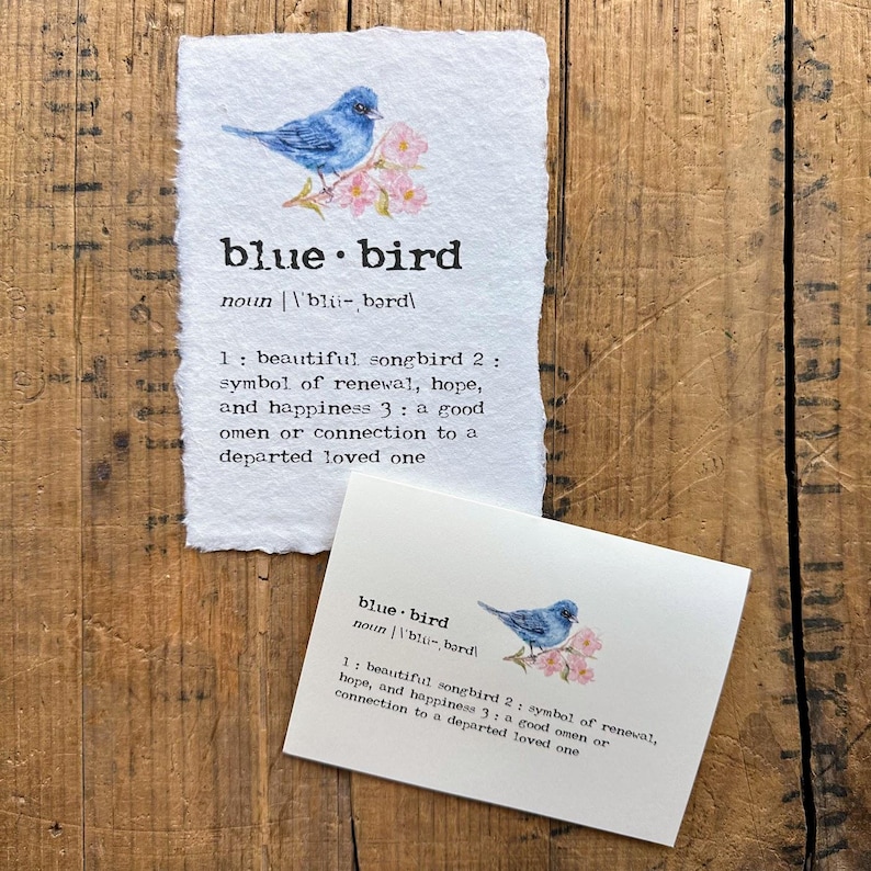 May include: A bluebird perched on a branch with pink flowers. The image is a definition card with the word "bluebird" and its meaning. The definition states that a bluebird is a beautiful songbird, a symbol of renewal, hope, and happiness, and a good omen or connection to a departed loved one.