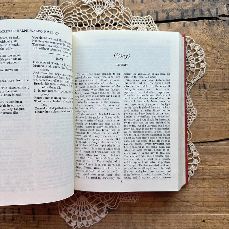 May include: A close-up of a book with a red cover and gold lettering. The title of the book is "Essays" and the author is Ralph Waldo Emerson. The text on the page discusses the concept of history as a reflection of the universal mind.