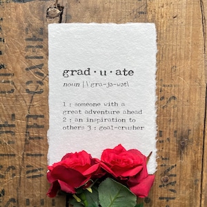 Peut inclure: Une carte blanche avec le mot "graduate" défini comme "quelqu'un avec une grande aventure à venir, une inspiration pour les autres, un briseur d'objectifs". La carte est posée sur une surface en bois avec deux roses rouges au premier plan.