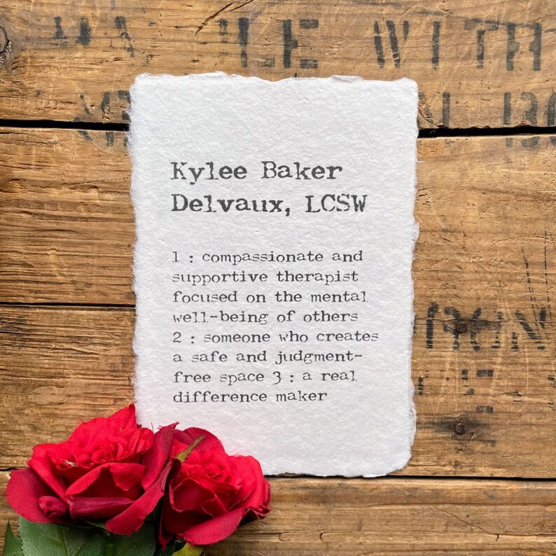 May include: A white card with black text on a wooden background. The text reads "Kylee Baker Delvaux, LCSW 1: compassionate and supportive therapist focused on the mental well-being of others 2: someone who creates a safe and judgment-free space 3: a real difference maker". Two red roses are in the bottom left corner of the image.